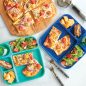 2 meal trays filled with pizza, salad, fruit, and bars with cut pizza on cutting board beside trays