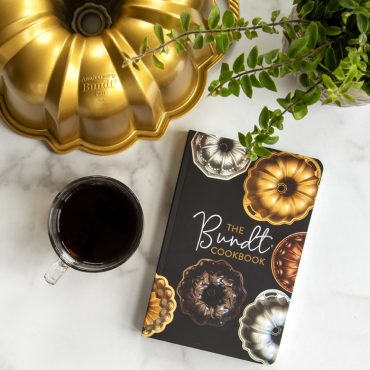 The BundtÂ® Cookbook with cup of coffee, Anniversary Bundt Pan and green plant on marble surface.