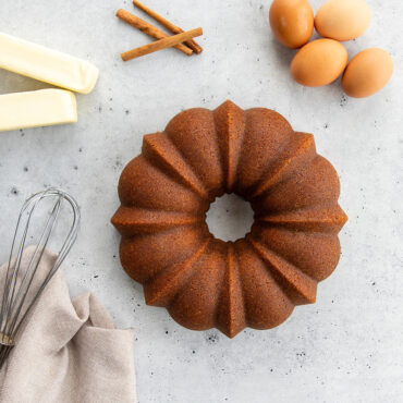 Golden brown cinnamon bundt cake in Nordic Ware fluted pan surrounded by eggs, butter, cinnamon sticks and whisk