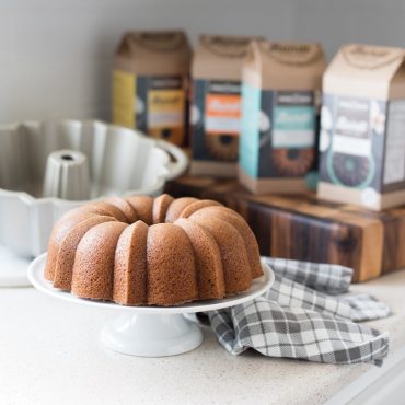 Baked Cinnamon Spice bundt cake on cake stand, Anniversary BundtÂ® cake and all four cake mixes in background