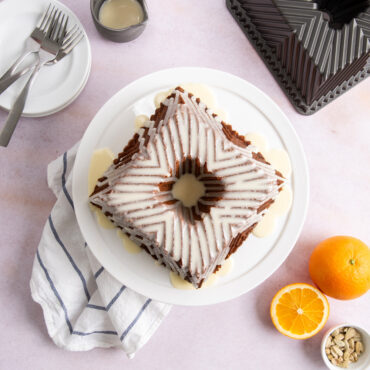 Glazed Bundt Squared cake on white cake stand against pink background, oranges next to plate