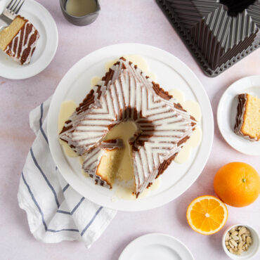 Glazed and cut Bundt Squared cake on white cake stand against pink background, oranges next to plate