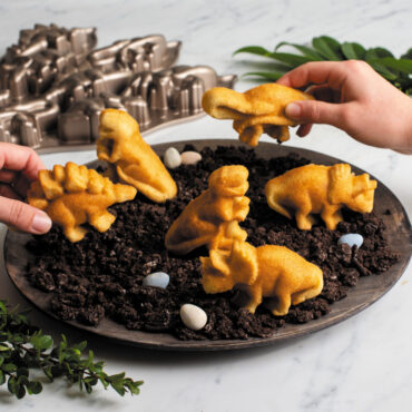 dinosaur cakelets on wooden plate with crushed cookies and candy dinosaur eggs, two hands holding two cakelets, with pan and green leaves