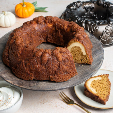 Autumn Wreath Bundt on wooden cake stand, piece cut out showing cream cheese filling with pan and mini pumpkins in backround
