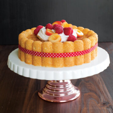 Whole baked cherry charlotte cake marbled, garnished with whipped cream and fresh raspberries, on cake stand