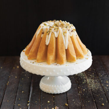 Golden bundt cake baked in Nordic Ware Lotus bundt pan with white glaze dripping down fluted sides, topped with chopped nuts on white cake stand