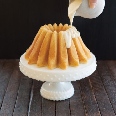 Golden bundt cake with lotus petal design on white cake stand, white glaze being poured from small pitcher