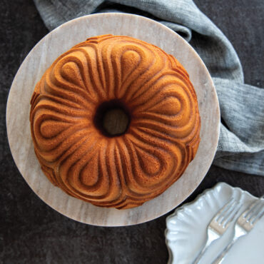 Golden brown chiffon bundt cake with decorative swirled ridges on white cake stand, baked in Nordic Ware pan