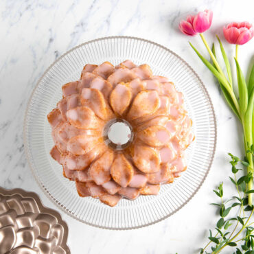 Overhead image of Blossom Bundt cake with pink frosting on clear cake stand with pink flowers and pan around