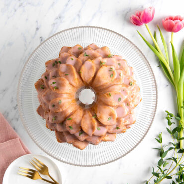 Overhead image of Blossom Bundt cake with pink frosting and green leaves sprinkled on clear cake stand with pink flowers and pan around