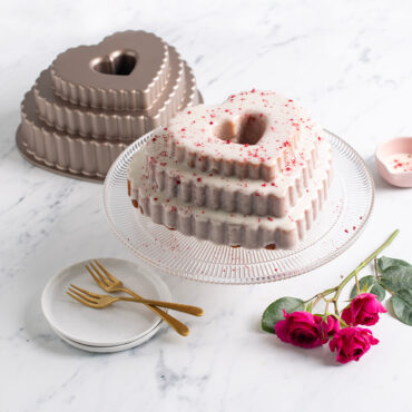 Tiered Heart Bundt with white frosting and red sprinkles on clear cake stand with roses and pan around it