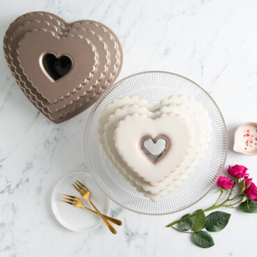 White frosted Tiered Heart Bundt on clear cake stand surrounded by roses and pan, overhead