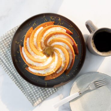 Golden bundt cake with white glaze on dark plate, Nordic Ware Heritage aluminum bundt pan and coffee cup nearby