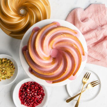 Golden bundt cake with pink glaze on white pedestal, Nordic Ware Heritage aluminum bundt pan and toppings nearby
