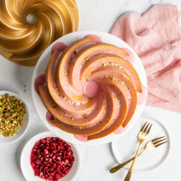 Golden bundt cake with pink glaze on white pedestal, Nordic Ware Heritage aluminum bundt pan and toppings nearby