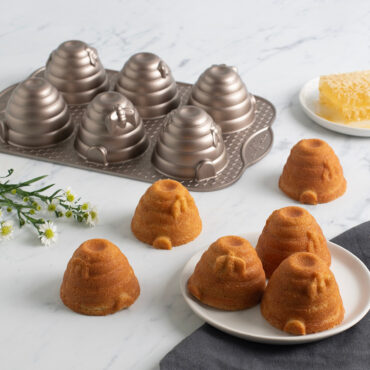 Golden-brown beehive cakelets with detailed ridged texture, four on white plate and one on marble surface with honey comb and flowers