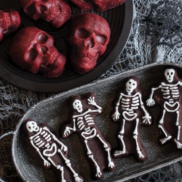 Baked chocolate skeleton cakelets with piped white frosting to show details, on serving plate, red velvet skull cakelets on plate beside skeleton cakes, web decorations