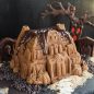 Baked Haunted Manor spice cake with thick chocolate glaze on roof, on counter with mini chocolate chips around house, slivered almonds and chopped almonds for walkway, tree with red lit up face in background