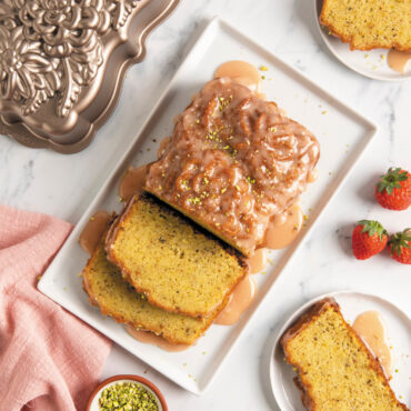 Frosted Wildflower loaf cake cut on rectangular plate with pan, strawberries, slices of cake, and pasticcios in a small dish