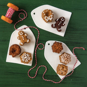 Assorted baked teacake cakelets in holiday shapes including Christmas tree, snowflake, and ornament designs on white plates