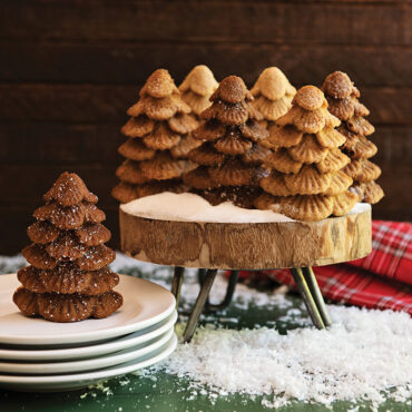 Four golden evergreen tree cakes standing upright on white cake stands in winter holiday scene