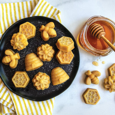 Golden bee-shaped bitelets arranged on black plate beside glass honey jar, showing detailed Nordic Ware pan results