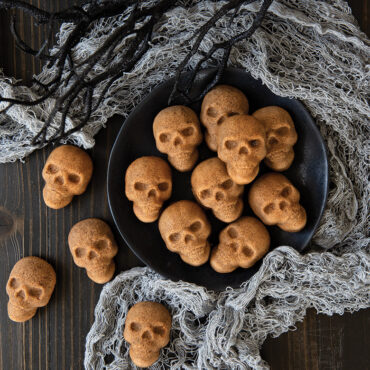 Small golden-brown skull-shaped cakelets arranged on white plate with Halloween decorations in background