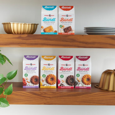Nordic Ware bundt cake mix collection displayed on wooden shelves with bronze bundt pan and baking dishes