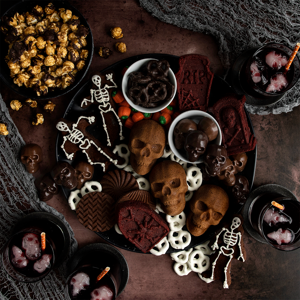 Halloween-themed charcuterie board with skull-shaped cakes, orange cheese, crackers, and seasonal decorations arranged on dark surface