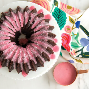 Baked chocolate Brilliance 5 Cup Bundt with pink glaze, container with glaze on the side with floral towel.