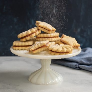 Stamped cookie sandwiches filled with apricot jam on cake platter, being dusted with powdered sugar