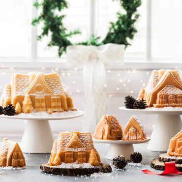 Three baked gingerbread house cakes in various sizes on white cake stands, dusted with powdered sugar