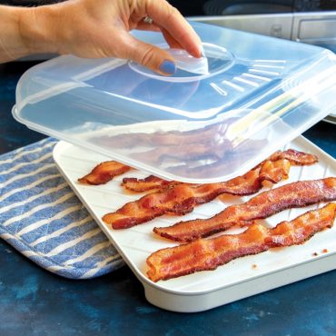 Hand pulling off lid from slanted tray with cooked bacon, microwave in background.
