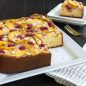 Baked peach and raspberry cake made in square springform pan, cut piece on plate in background.