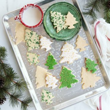 Baked Christmas tree shaped cut out cookies on embossed tree baking sheet with a glass of milk on sheet pan tray, towel