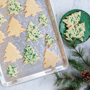 Plain and sprinkled cut out tree cookies on embossed tree baking sheet, plate with cookies next to sheet pan.