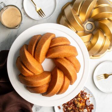 Golden glazed bundt cake with braided rope design next to Nordic Ware aluminum bundt pan and serving plate