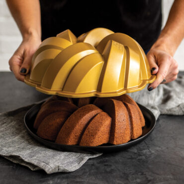 Hands holding Nordic Ware 75th Anniversary aluminum bundt pan above golden baked cake with braided pattern