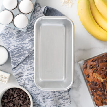 Nordic Ware natural aluminum 1-pound loaf pan with baking ingredients including flour, eggs, and butter arranged around it