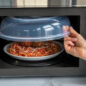 Hand taking off spatter cover from bracket  to place on top of plate of food in microwave