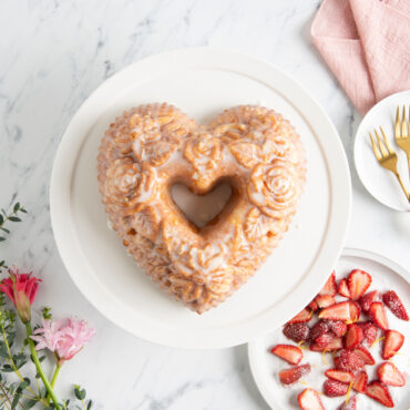 Overhead of glazed Floral Heart Bundt on white cake stand with a plate of diced strawberries and flowers