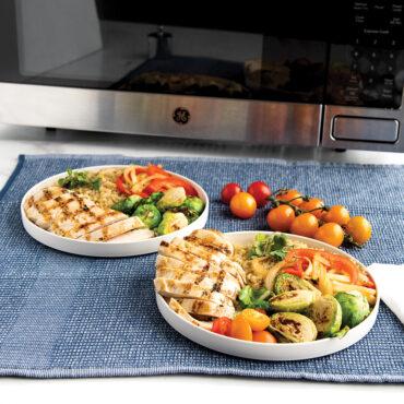A white lunch plate on a blue placemat sitting on a white table. The plate contains a serving of food. In the background there is a microwave showing that these plates are microwave safe.