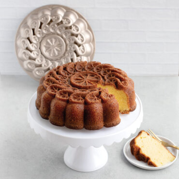 A citrus twist cake on a white cake stand,  with a golden brown piece of the cake sliced and ready to eat on a white plate next to the citrus cake that's on the cake stand.