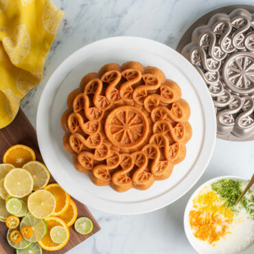 citrus twist cake on white cake stand with sliced lemons, pan, and bowl of glaze