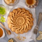 A delicious citrus twist cake on a surface overhead. The cake has a light golden brown crust and honey and cake ingredients are around the cake with a fork.