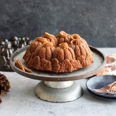 The image showcases a side angle of a baked pumpkin patch bundt cake on a cake stand with the pan and plates in background.