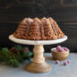 The image showcases a beautifully baked Very Merry Bundt cake displayed on a cake stand adorned with festive pine leaves and vibrant cranberries.