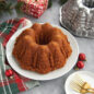 The image showcases a beautifully baked Very Merry Bundt cake displayed on a plate with the pan and present in background scene.