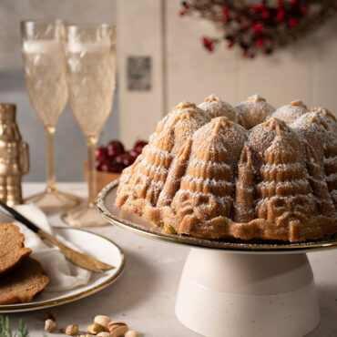 The image depicts a beautifully baked Very Merry Bundt cake, adorned with a delicate dusting of powdered sugar on its surface. In the background, a glass of champagne adds a touch of elegance.
