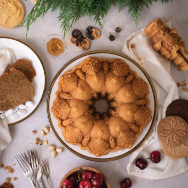 The image shows a visually appealing overhead shot of a beautifully baked Very Merry Bundt cake adorned with pine leaves, cherries, and other festive baked goods.
